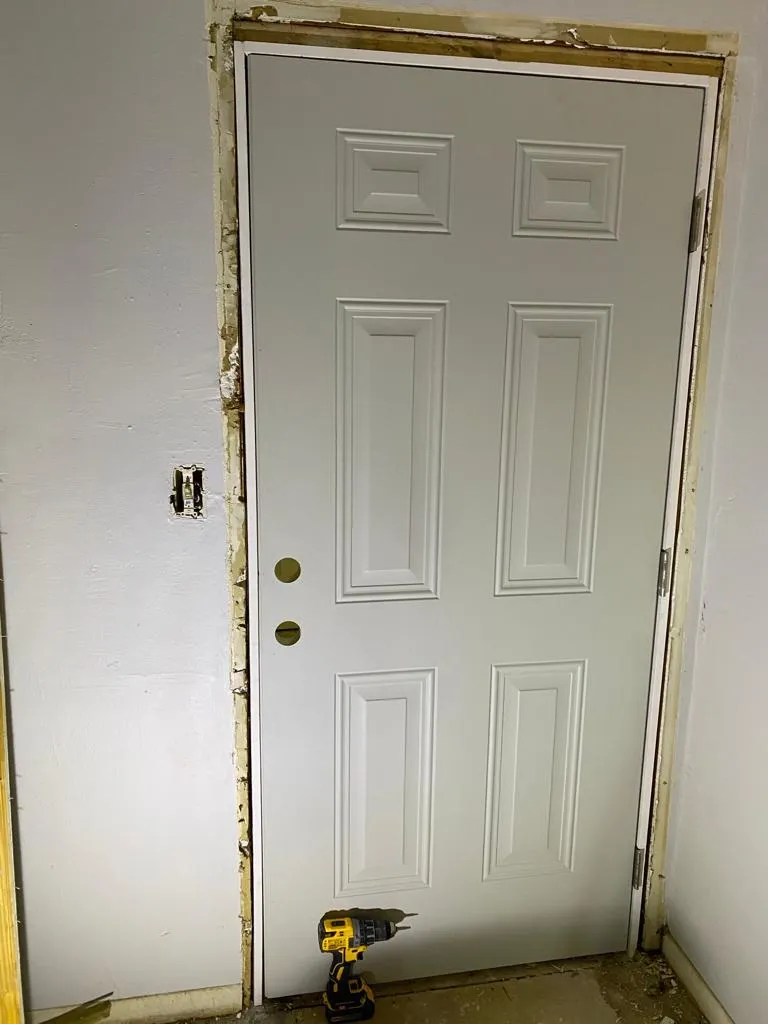 New six-panel door hung in a stripped frame by Family Solutions LLC, drill at its foot